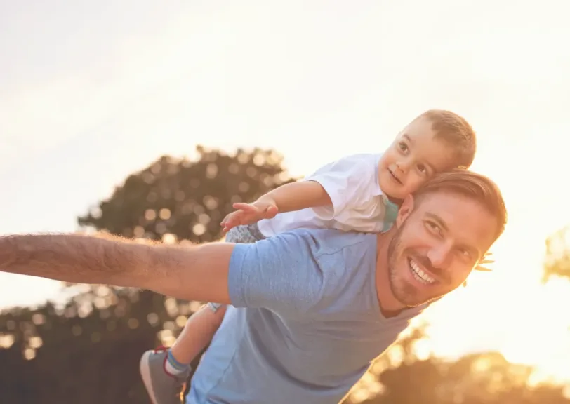A father is leaning over with his child on his back in a running forward monition. Both are smiling.