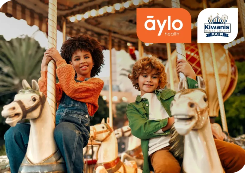 Aylo Health and Kiwanis Henry County Fair logos are on top of a picture of two children smiling while on a carousel