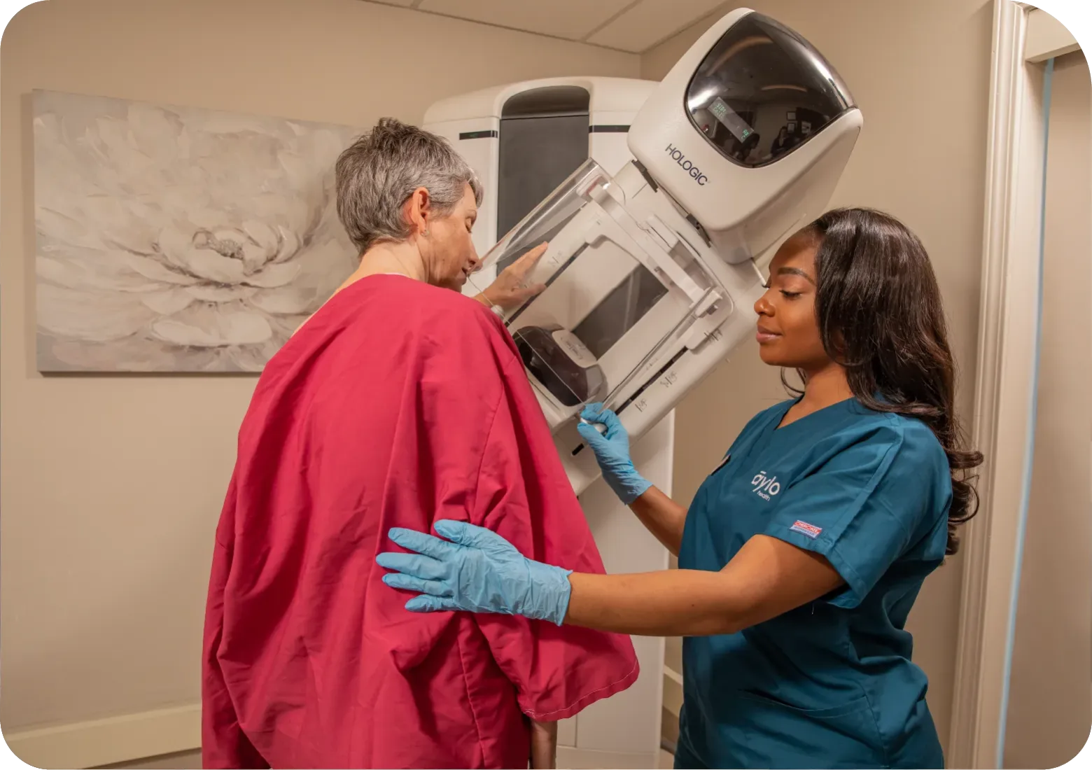 An Aylo nurse is standing next to a patient who is getting her mammogram.